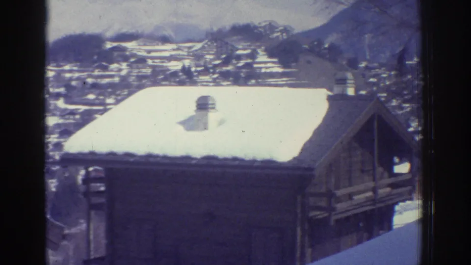 Late 1970s alpine village scene on 8mm film: snow-covered wooden rooftops with a metal chimney vent, dark fir trees, and mountain terrain; consistent with a Swiss highland location.