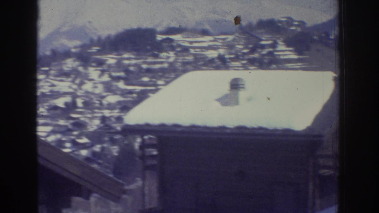 Snow-covered wooden chalet rooftop in foreground with an Alpine village and mountains behind, filmed on 8mm.