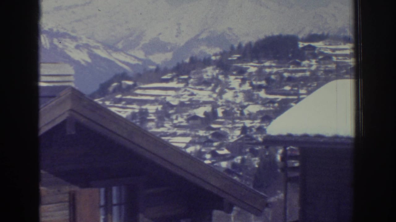 8mm film frame c.1976 — gable end of snow-covered chalet with an Alpine village and mountain ridgeline beyond, likely Swiss Alps.