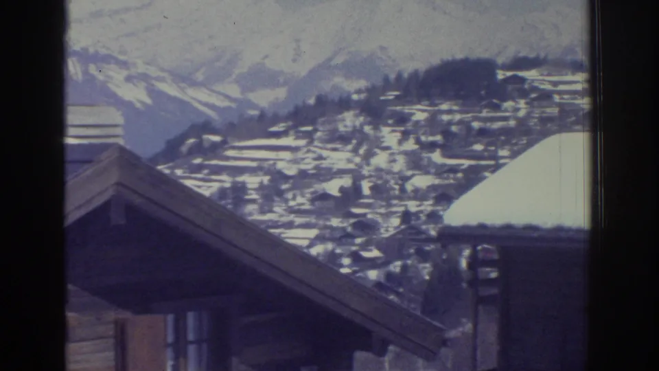 8mm film frame c.1976 — gable end of snow-covered chalet with an Alpine village and mountain ridgeline beyond, likely Swiss Alps.