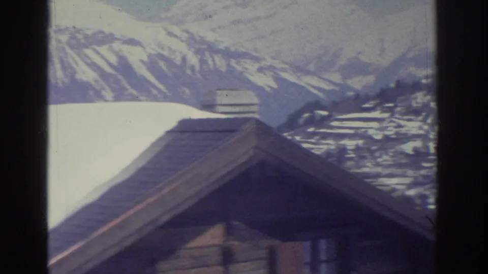 8mm home movie frame c.1976 — wooden chalet roof at the bottom with a large snow-capped Alpine mountain ridge dominating the upper frame, likely Swiss Alps.