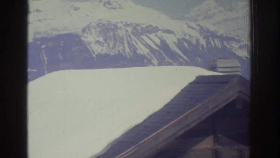 8mm film frame c.1976 — two adjacent chalet rooftops under heavy snow with an Alpine mountain range in background, likely Swiss Alps.