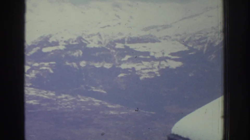 Wide alpine panorama of snow-covered peaks filmed through a vehicle window, likely Swiss Alps region circa 1976, with 8mm grain and blue-purple color shift.