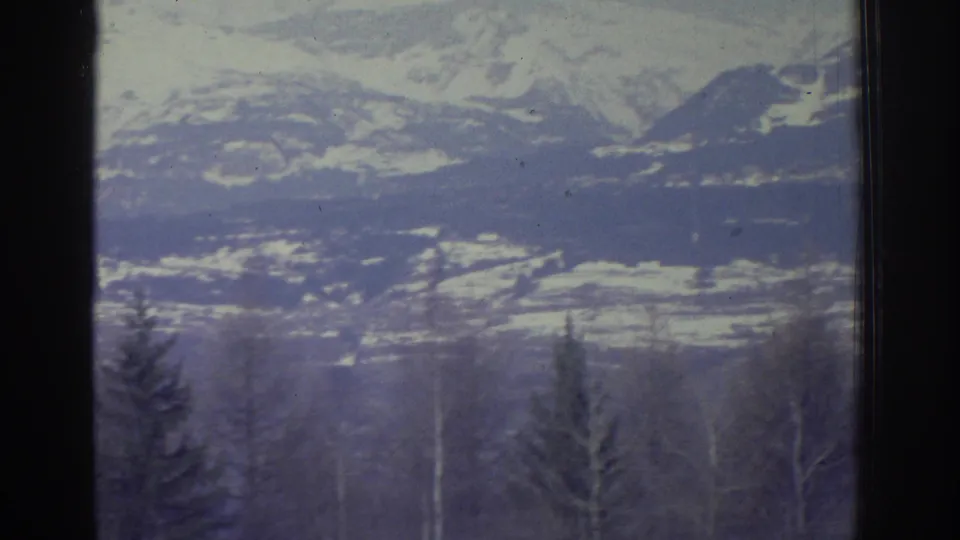 8mm home movie frame from 1976 showing a snow-covered Alpine ridgeline with coniferous trees in the foreground and a pale overcast sky above.