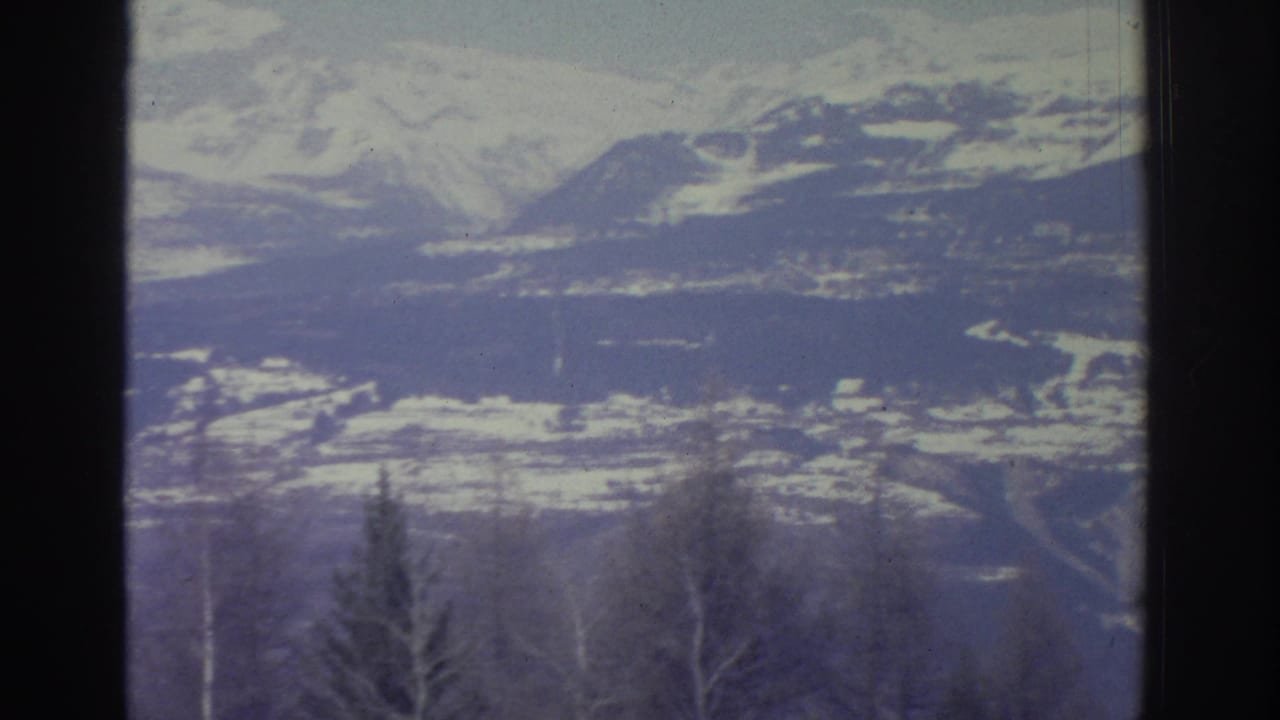 8mm home movie frame from 1976 showing a wide Alpine winter panorama with a snowy valley basin, layered ridgelines, and sparse foreground trees.