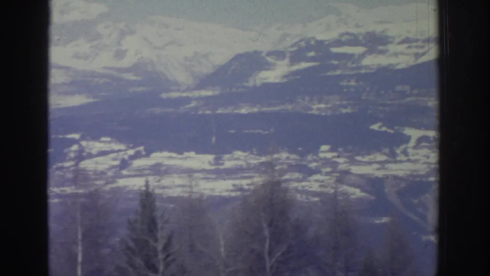 8mm home movie frame from 1976 showing a wide Alpine winter panorama with a snowy valley basin, layered ridgelines, and sparse foreground trees.