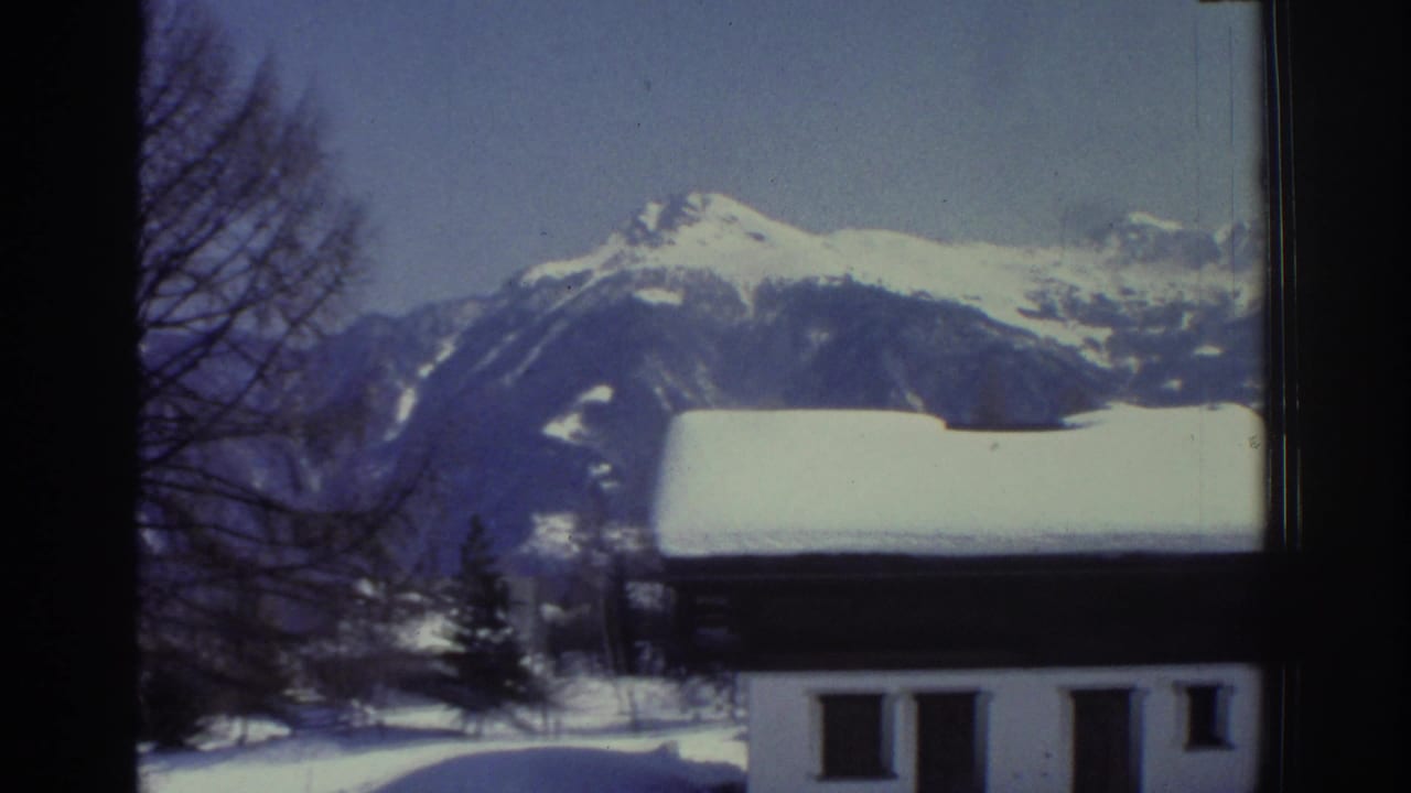 Snow-capped mountains behind a white building with dark windows, bare trees at right, winter alpine scene on 8mm film.