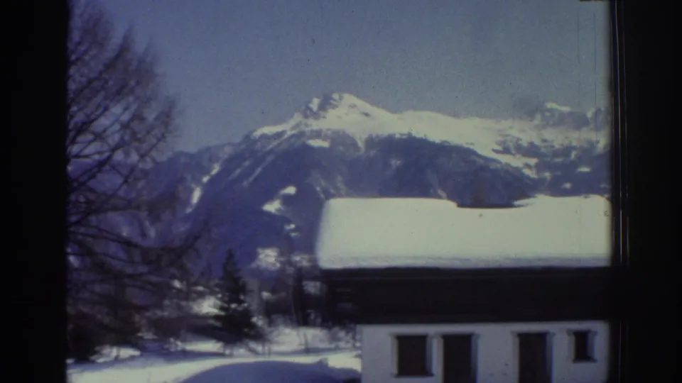 Swiss Alps winter scene on 8mm, 1976: white low-rise building below snow-capped peaks, bare trees at right, cool blue 1970s color palette.