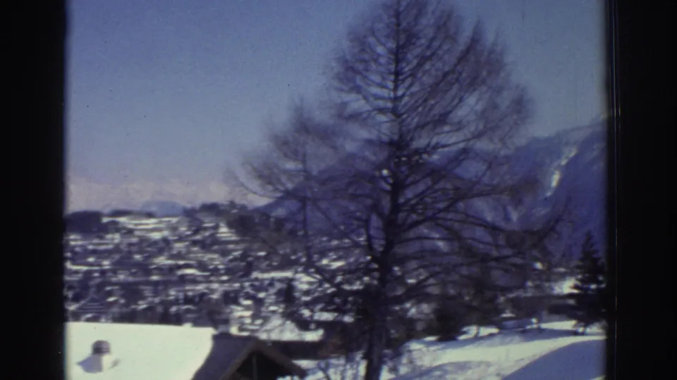 Switzerland, 1976, 8mm: panoramic elevated view of a snow-covered Alpine valley town, bare tree and conifers in foreground, slopes stretching to the horizon.