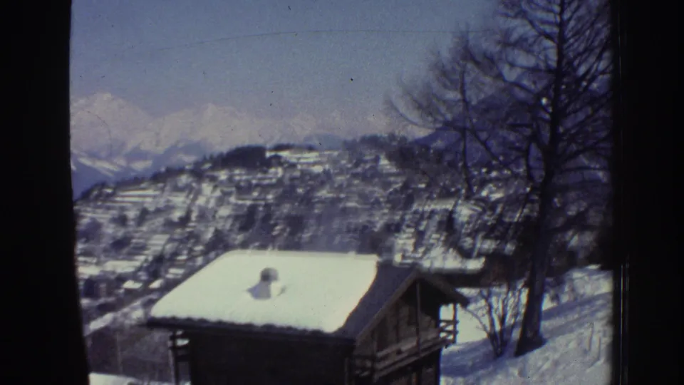 Switzerland, 1976, 8mm: traditional cabin's snow-covered roof in foreground with terraced hillside and alpine peaks receding behind, bare tree at right.