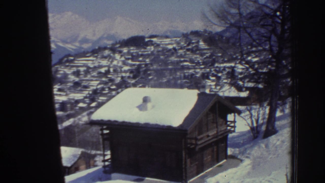 Snow-covered wooden alpine chalet in foreground with hillside village and mountain peaks behind, 8mm film border visible on left edge.