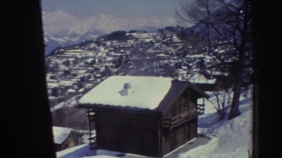 8mm home movie frame from 1976 showing a snow-laden wooden chalet with a hillside Swiss village and Alpine peaks visible in the background.
