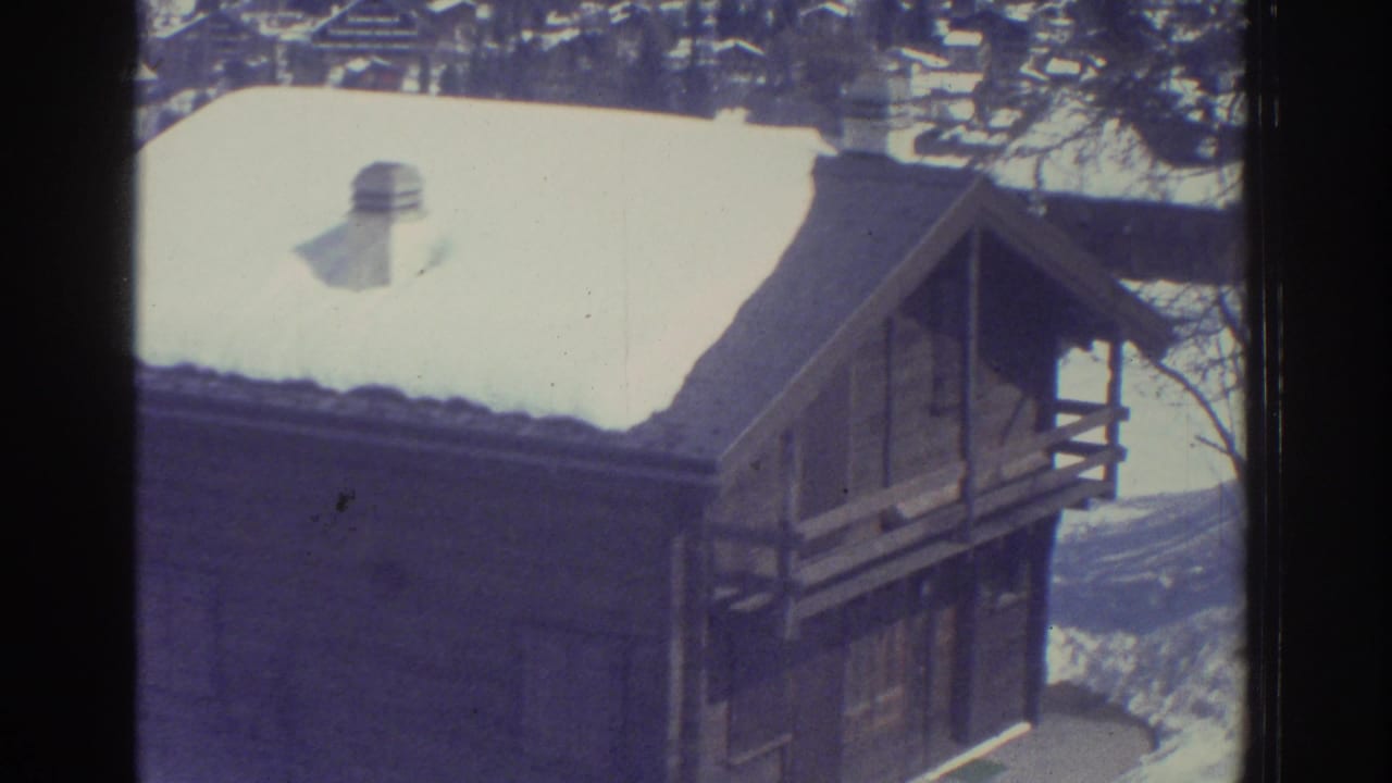 Close-up of a wooden alpine chalet with steep snow-covered pitched roof, metal vent cap, and wooden balcony railing.