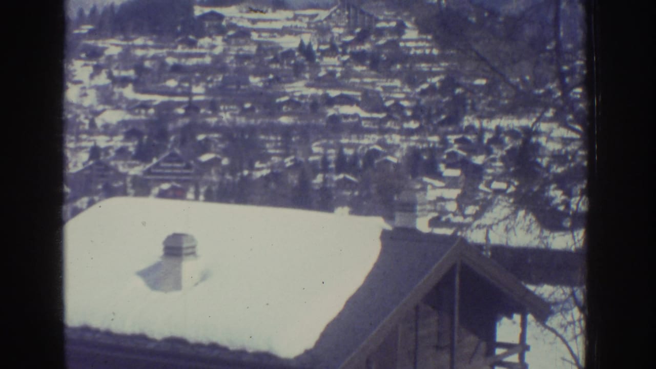 8mm frame from 1976 showing a foreground chalet roof and a dense snow-covered Alpine village spreading across a hillside, consistent with the Bern region.