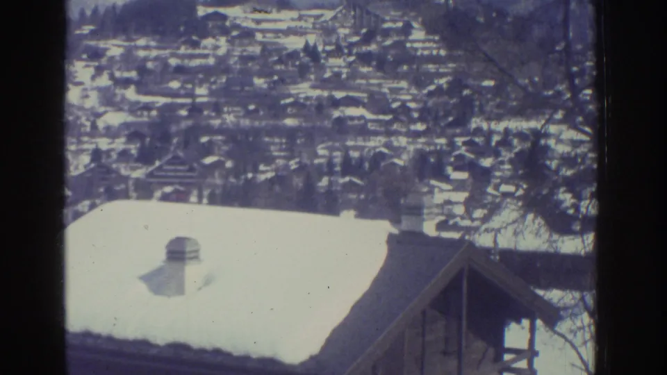8mm frame from 1976 showing a foreground chalet roof and a dense snow-covered Alpine village spreading across a hillside, consistent with the Bern region.