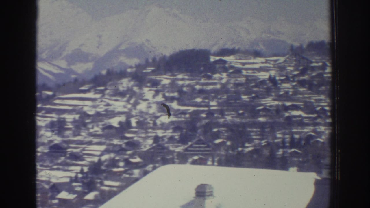 8mm home movie frame from 1976 showing a panoramic winter Alpine landscape with a snow-covered village on a hillside and mountain peaks in the distance.