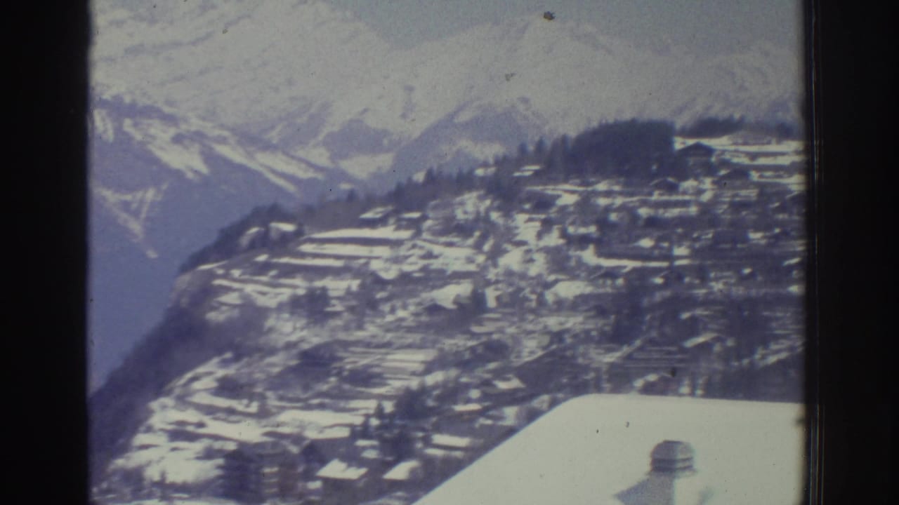 Snow-covered alpine village on terraced slopes viewed from above, with mountain ridges in the background, 8mm film.