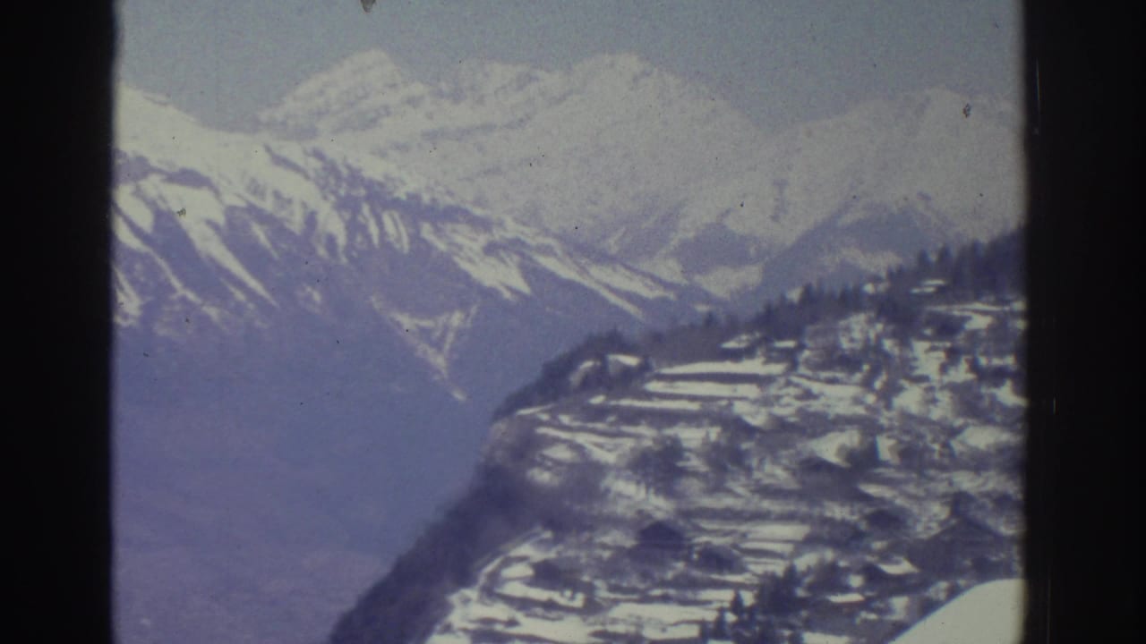 Snow-covered mountain ridges dominating the frame mid-pan, with scattered buildings visible in the lower corner, 8mm film.