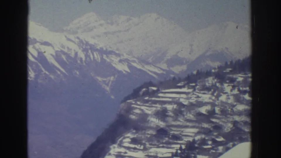 8mm pan frame showing snow-covered Alpine mountain ridges and scattered buildings below, possibly Swiss Alps region, circa 1976.