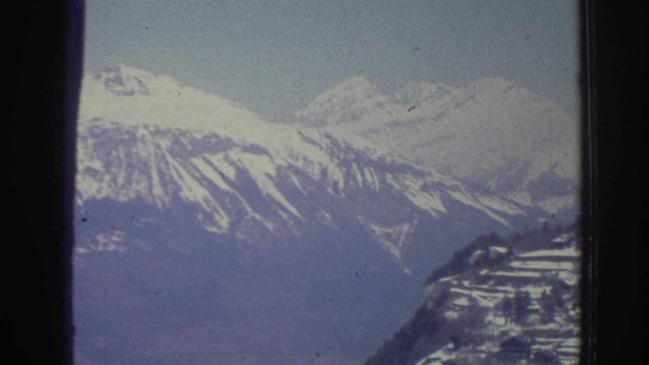 Wide 8mm view of snow-covered Alpine slopes and distant valley settlement below, possibly Swiss Alps region, circa 1976.