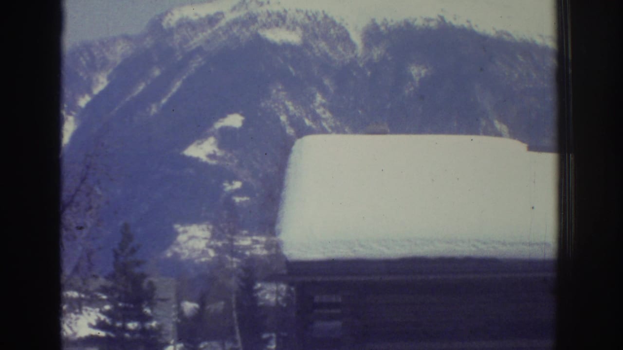 Snow-covered rooftop in close foreground against an alpine mountain slope and pine tree, possibly Swiss Alps region, circa 1976, 8mm film.