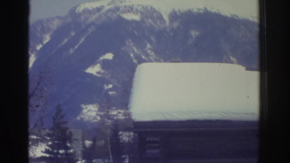 Snow-covered rooftop in close foreground against an alpine mountain slope and pine tree, possibly Swiss Alps region, circa 1976, 8mm film.