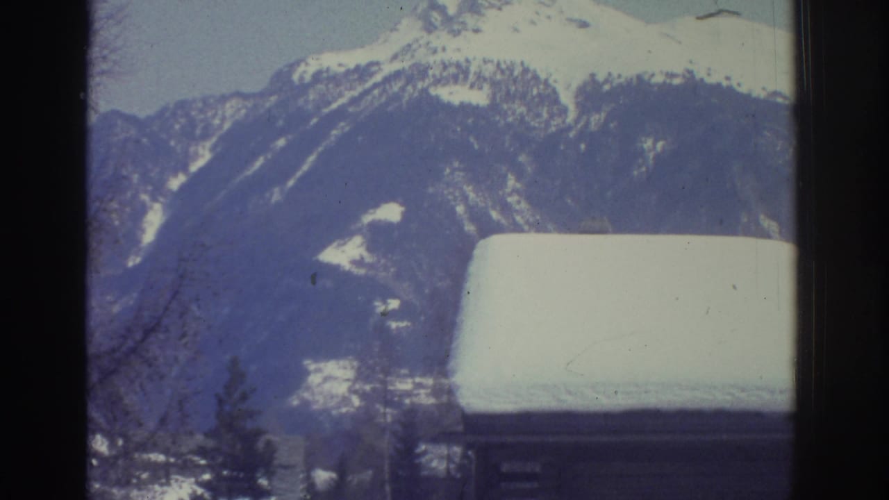 Snow-covered alpine mountain peak rising above a snow-laden chalet rooftop in the foreground, with conifer trees at left.