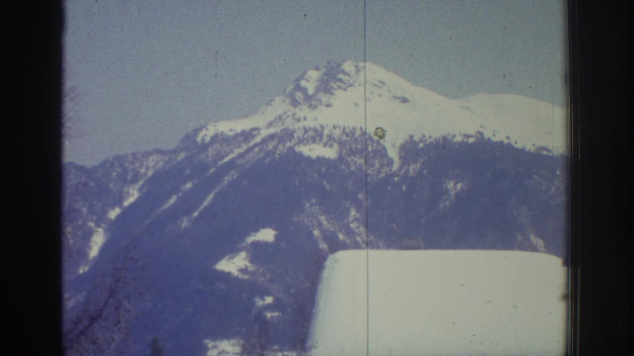 Alpine mountain peak centered under clear pale blue sky, with a snow-covered chalet rooftop below and a small green film artifact visible on the ridge.