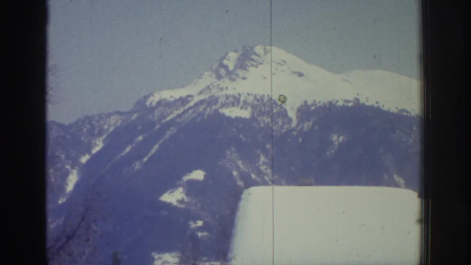 8mm home movie frame, likely Switzerland mid-1970s, showing an alpine peak under clear sky with a snow-covered rooftop below; a small green lens artifact is visible on the ridgeline.
