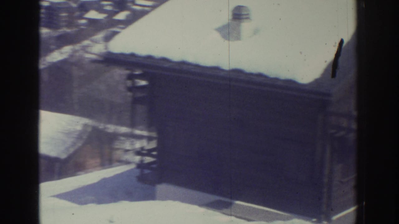 8mm home movie frame, likely Swiss Alps mid-1970s, showing a close view of a timber chalet rooftop bearing heavy snow, with a chimney vent and adjacent building rooftops visible below.