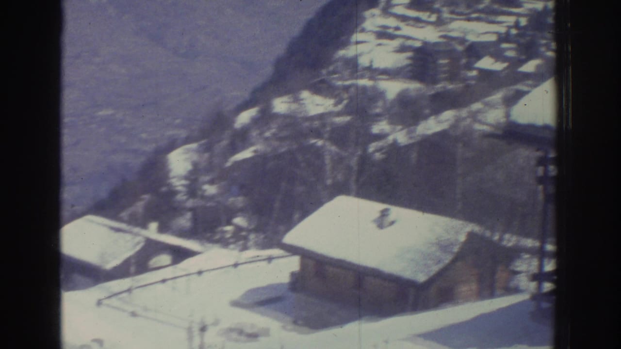 8mm home movie frame, likely Swiss Alps mid-1970s, showing an elevated perspective on a small alpine village with several timber chalets under deep snow, utility poles visible at right.