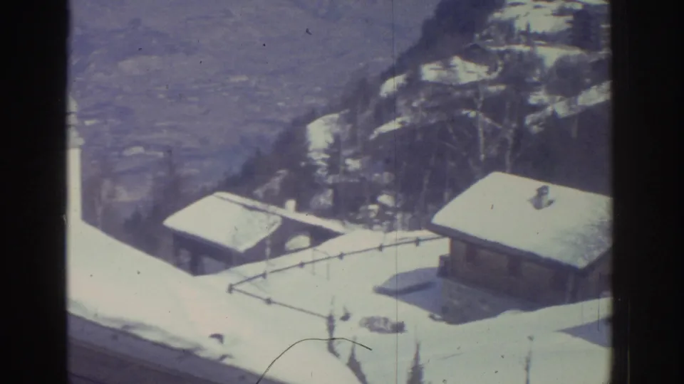 Elevated 8mm shot looking down onto snow-laden pitched rooftops of alpine-style buildings with valley and mountain slope behind; attributed to Bern, Switzerland, 1976.
