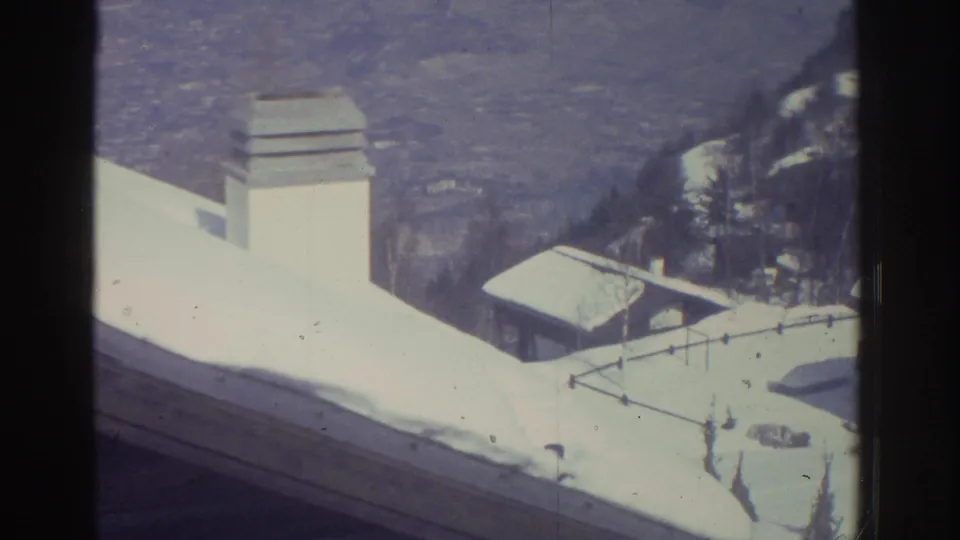 8mm home-movie view from a rooftop looking across snow-laden slopes and a chimney stack toward lower alpine buildings and valley; attributed to Bern, Switzerland, 1976.