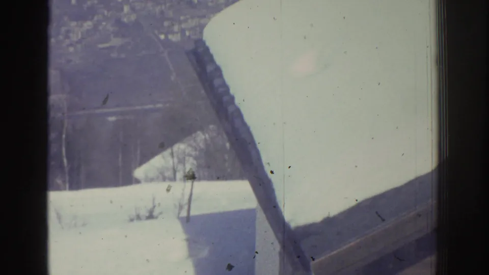 8mm pan continuing from rooftop vantage showing a broad snow-covered roof in foreground, utility pole, and snow-covered settlement below; attributed to Bern, Switzerland, 1976.