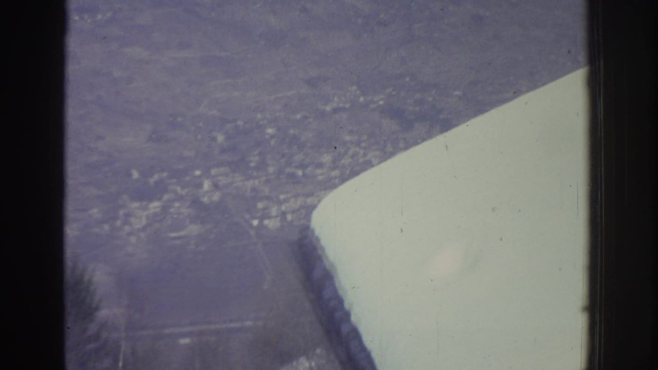 Aerial view through an airplane window of a snow-covered city far below, with the aircraft wing filling the lower foreground.