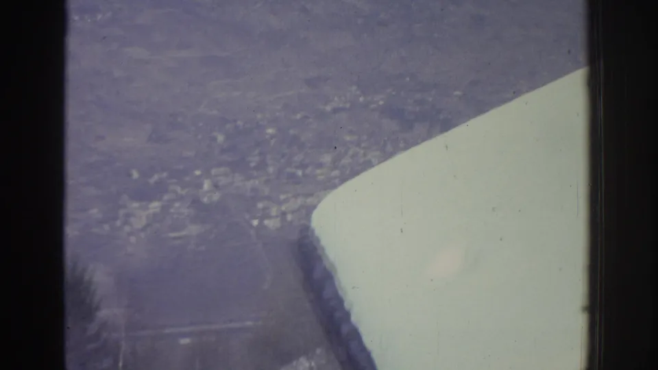 8mm aerial shot circa 1976 looking down through an airplane window at a snow-covered Swiss city, wing prominent in the lower foreground.