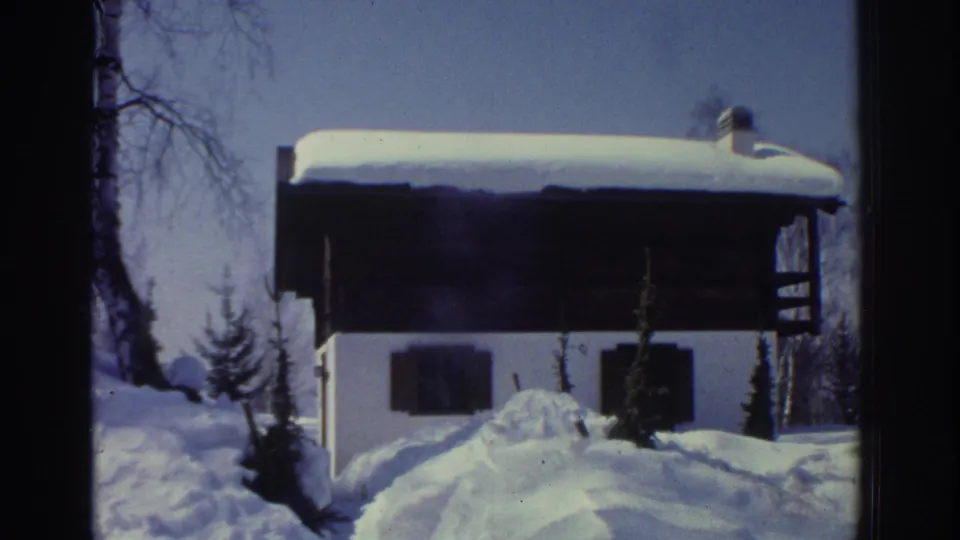 8mm home movie circa 1976 showing traditional Swiss chalet exterior: white walls, dark timber upper level, chimney, deep snowdrifts in foreground, clear blue sky.