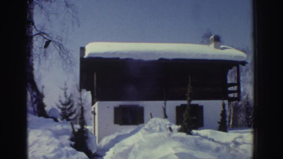 8mm footage circa 1976 of traditional Swiss chalet: white walls, dark timber balcony railing, chimney, heavy snow on roof, conifer and bare branches at sides.