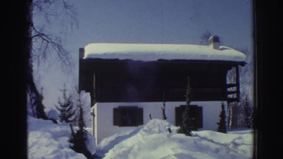 1970s 8mm home movie frame showing a white-walled Swiss alpine chalet with dark shutters under heavy snow, bare trees and conifers against a pale blue sky, circa 1976.