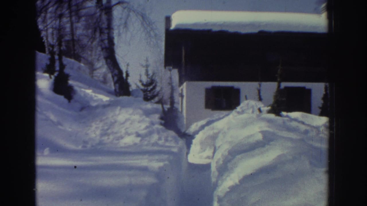 1970s 8mm home movie frame showing a narrow shoveled path cut through deep snow with steep snowbanks, an alpine chalet partially visible in the background, circa 1976.