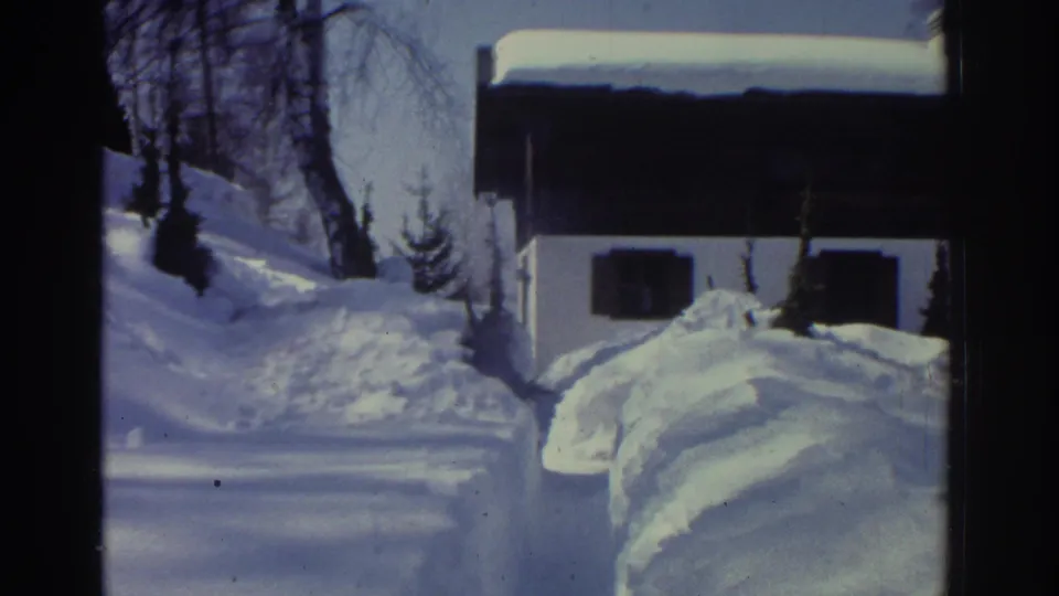 1970s 8mm home movie frame showing a narrow shoveled path cut through deep snow with steep snowbanks, an alpine chalet partially visible in the background, circa 1976.