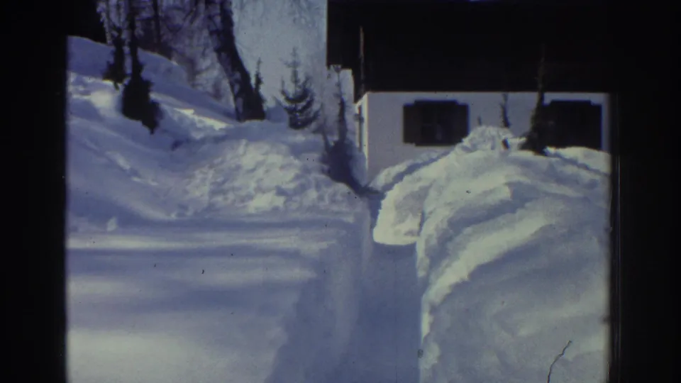 1970s 8mm home movie frame showing a panned-back view of the snowy alpine property, with the shoveled path in the foreground and a white chalet among conifers under a pale sky, circa 1976.