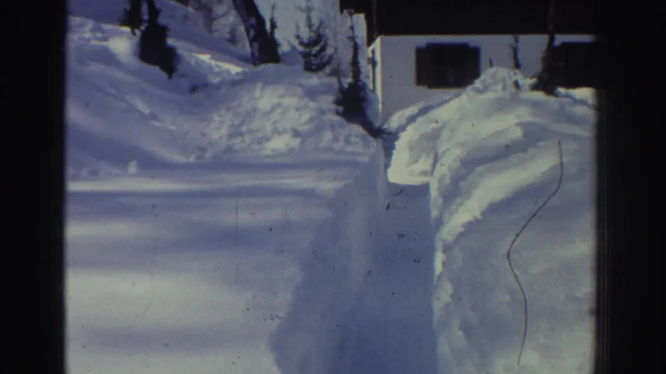 1970s 8mm home movie frame showing a close view of the shoveled path between tall snowbanks, chalet at right edge; a visible film scratch or hair mark adds archival texture, circa 1976.