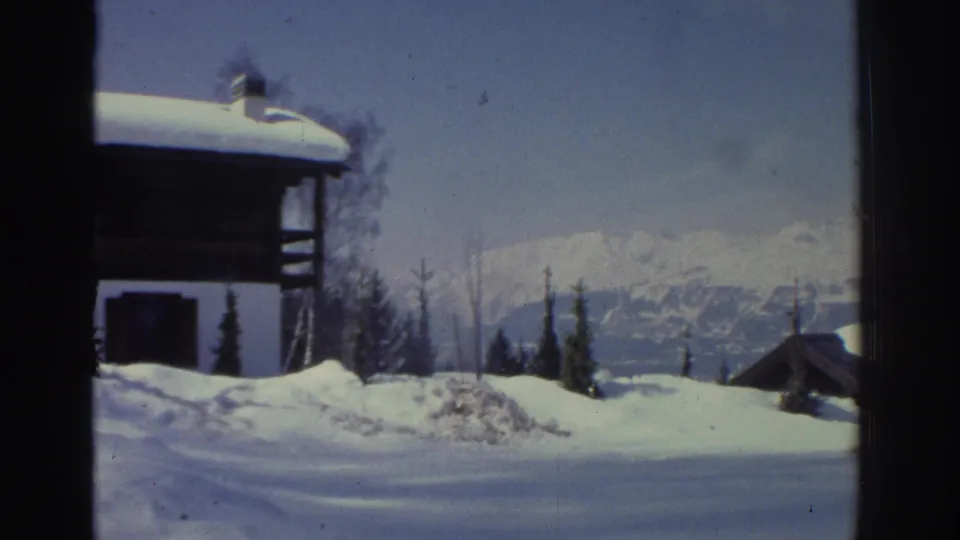 8mm home movie frame circa 1976 showing a timber chalet with snow-laden roof in the Swiss Alps, with conifers and distant peaks beyond.