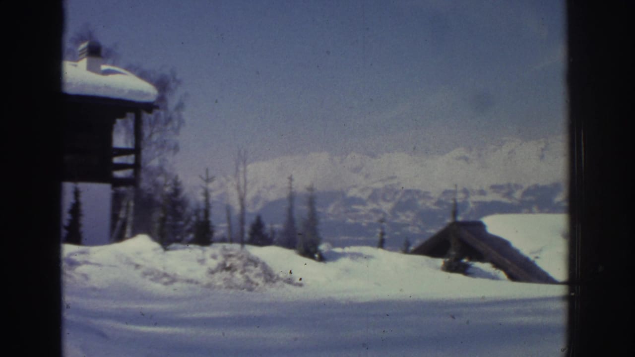8mm frame circa 1976 showing a timber chalet chimney and roof edge at upper right, with Swiss alpine winter landscape and distant peaks.