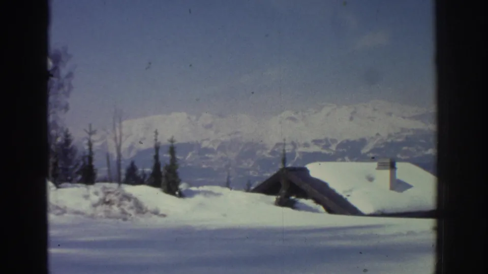 8mm home movie frame circa 1976 panning to reveal a broad Swiss alpine winter vista with a small snow-laden chalet, conifers, and far mountain peaks.