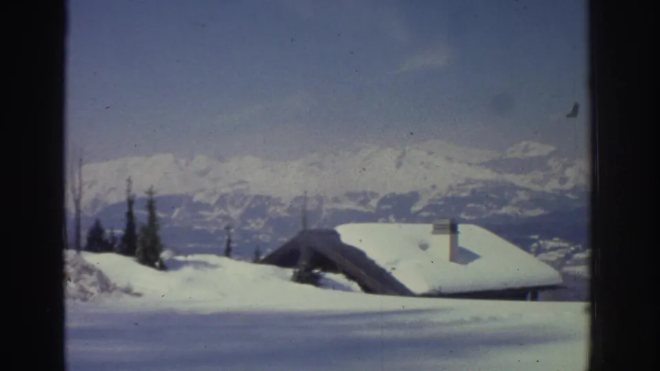 8mm frame circa 1976 showing a snow-laden chalet rooftop and chimney in the Swiss Alps, with conifers and a distant mountain ridge at the horizon.