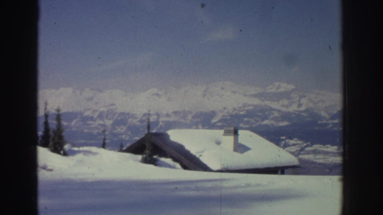 Snow-laden wooden chalets with chimneys set against a panoramic Swiss Alps ridgeline, circa 1976, 8mm home-movie footage.