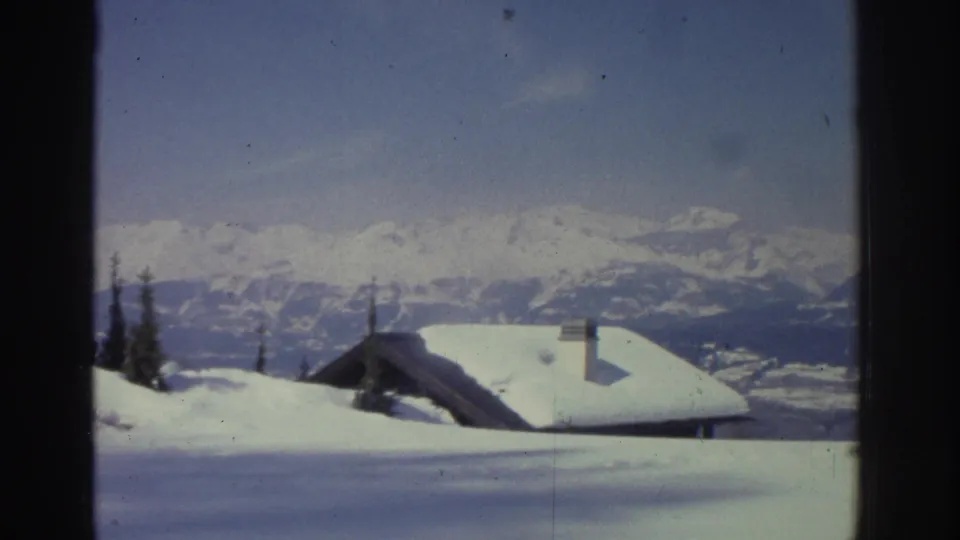 Snow-laden wooden chalets with chimneys set against a panoramic Swiss Alps ridgeline, circa 1976, 8mm home-movie footage.