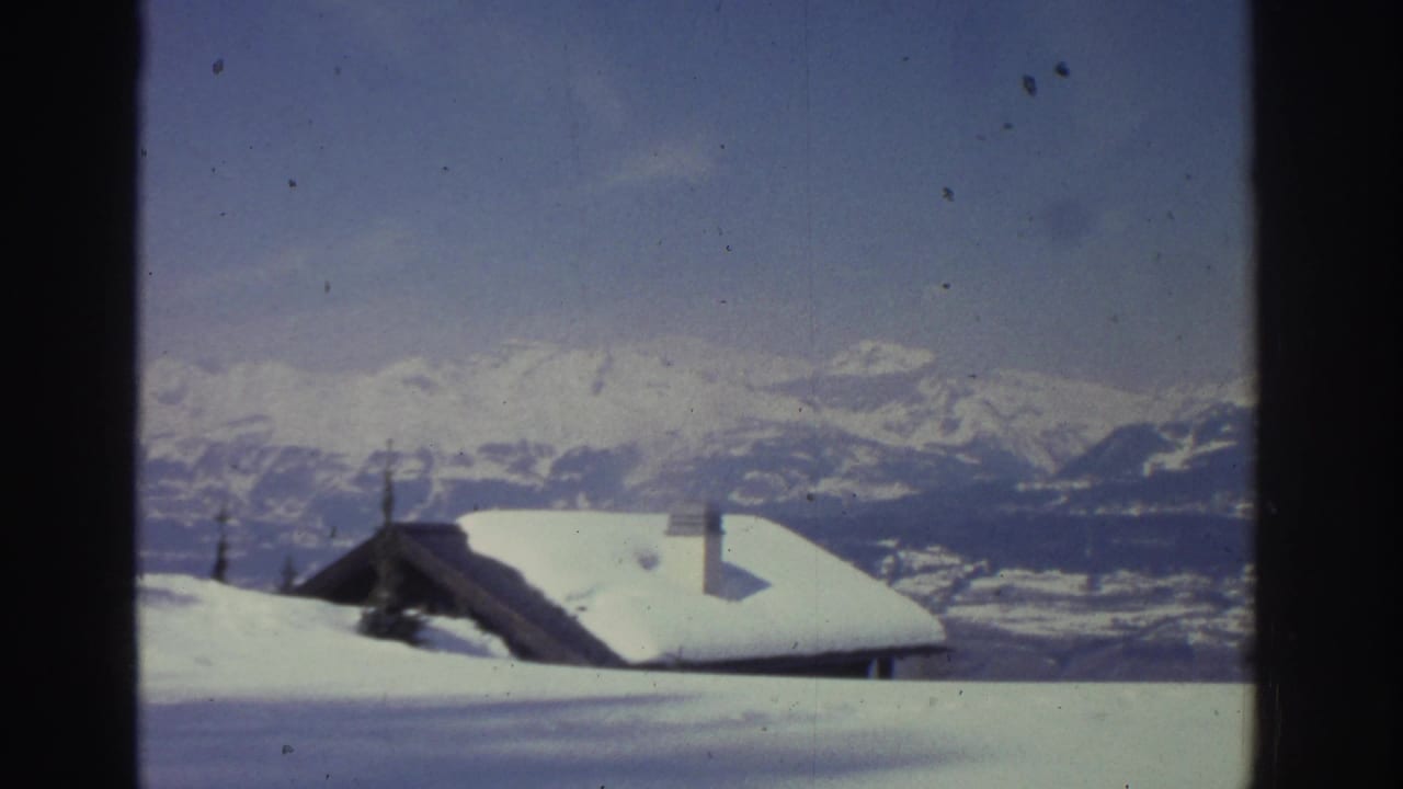 Continued view of two snow-laden alpine structures against a broad Swiss mountain ridgeline, mid-1970s 8mm footage.